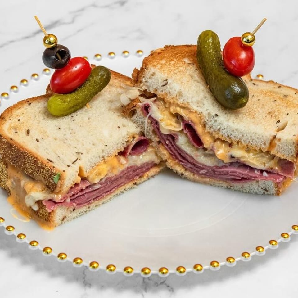 A toasted pastrami sandwich, cut in half, with melted cheese and sauce, garnished with pickles and tomatoes.