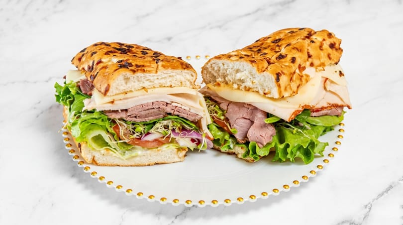 Two halves of a deli sandwich with roast beef, turkey, cheese, lettuce, and sprouts on a white plate.