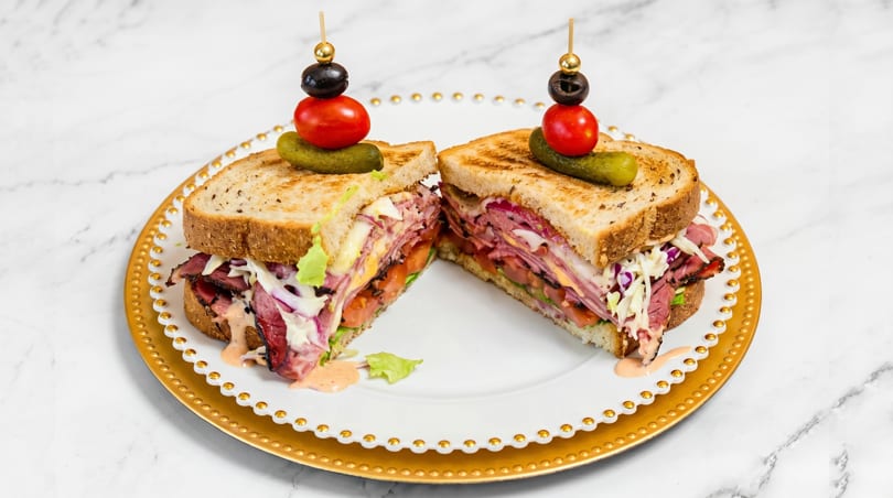 Two halves of a deli sandwich with pastrami, coleslaw, and sauce, garnished with a pickle and skewer.
