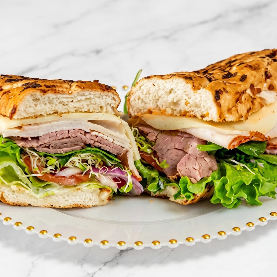 A large deli sandwich, cut in half, filled with meats, cheese, lettuce, and sprouts on a white plate.